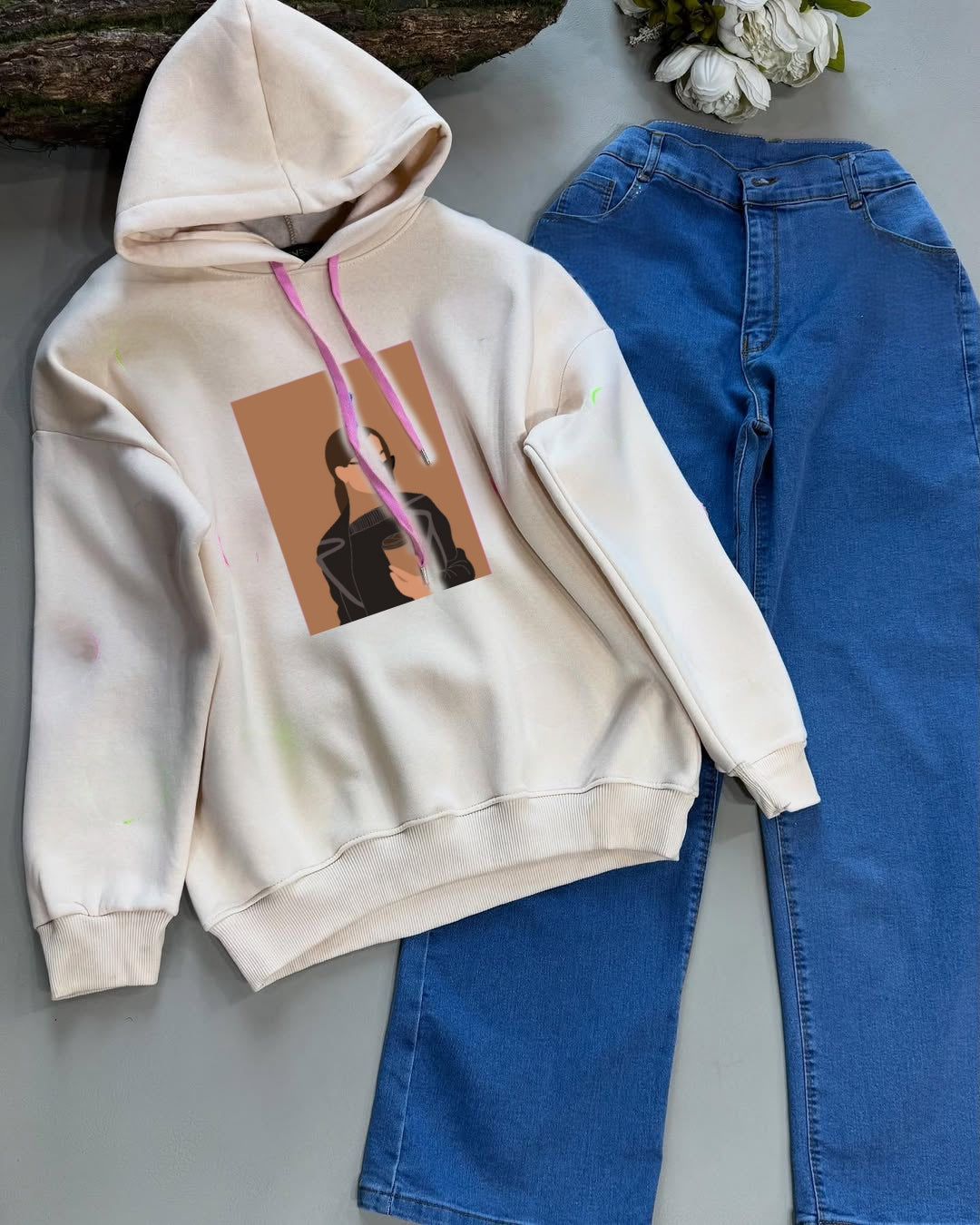 Beige hoodie with graphic design and blue jeans on a gray surface